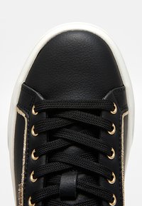 Baskets en cuir noir avec semelle blanche, lacets noirs et œillets dorés, vues de dessus en gros plan.