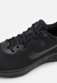 Chaussure de sport noire avec une tige en mesh, des accents synthétiques lisses et un logo de marque visible. Présente des lacets plats et une semelle rembourrée.