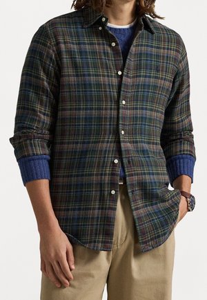 Homme portant une chemise à carreaux verte et bleu marine par-dessus un pull bleu, un pantalon beige et une montre en cuir marron au poignet gauche.