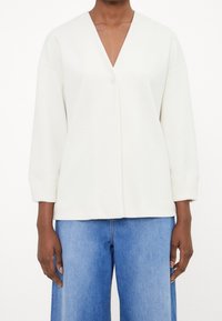 Vit långärmad topp med djup V-ringning och enkel knappstängning, med mjuk textur och lös passform. Matchas med blå vida jeans.