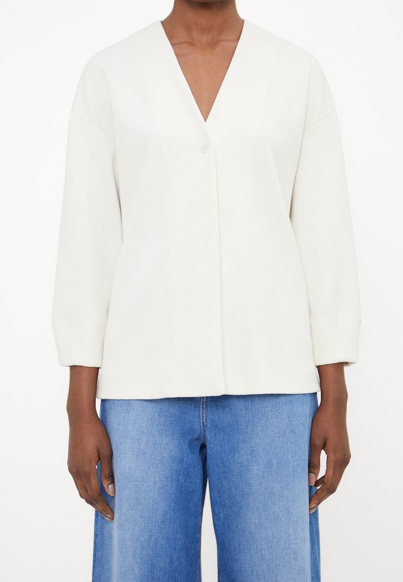 Vit långärmad topp med djup V-ringning och enkel knappstängning, med mjuk textur och lös passform. Matchas med blå vida jeans.