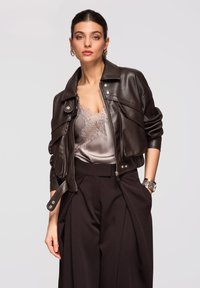 Femme portant une veste en cuir marron foncé, un caraco en satin bordé de dentelle, un pantalon noir taille haute et des bijoux en or sur un fond uni.