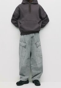 Sweat à capuche oversize gris foncé avec poche avant, associé à un pantalon cargo gris clair avec plusieurs poches et une coupe décontractée. Chaussures noires.