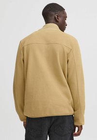 Beige fleece jacket with a relaxed fit, featuring a classic collar, long sleeves, and smooth texture, paired with dark pants.