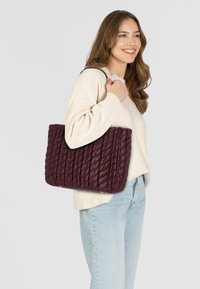 Sac en toile matelassé bordeaux avec une finition brillante, de forme rectangulaire et de pieds métalliques discrets, tenu par une personne portant un léger pull et un jean.