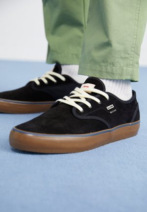 Black suede sneakers with white laces and brown soles worn with white socks and green pants on light blue floor.