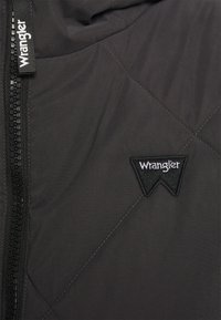 Czarna pikowana kurtka z zamkiem błyskawicznym, kontrastowym przeszyciem i czarną łatą w kształcie trójkąta z logo "Wrangler". Gładka tekstura.