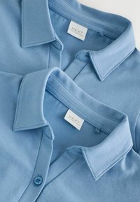 Svetlomodré polokošele s goliermi. Sú vybavené zapínaním na gombíky a majú hladkú textúru. Štítky označujú značku "NEXT" a uvádzajú, že sú vyrobené v Bangladéši.