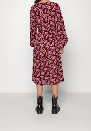 Kobieta w bordowej sukience z długim rękawem, sięgającej do kolan, z różowym kwiatowym wzorem i czarnych botkach za kostkę, stoi twarzą do białej ściany.