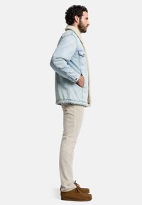 Ljusblå denimjacka med en beige faux fur-krage, bärs över krämfärgade byxor och bruna mockaskor, sedd från sidan.