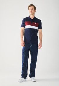 Menns polo-skjorte i marineblått med røde og hvite horisontale striper; har korte ermer, krage og brodert logo. Brukt med blå jeans.