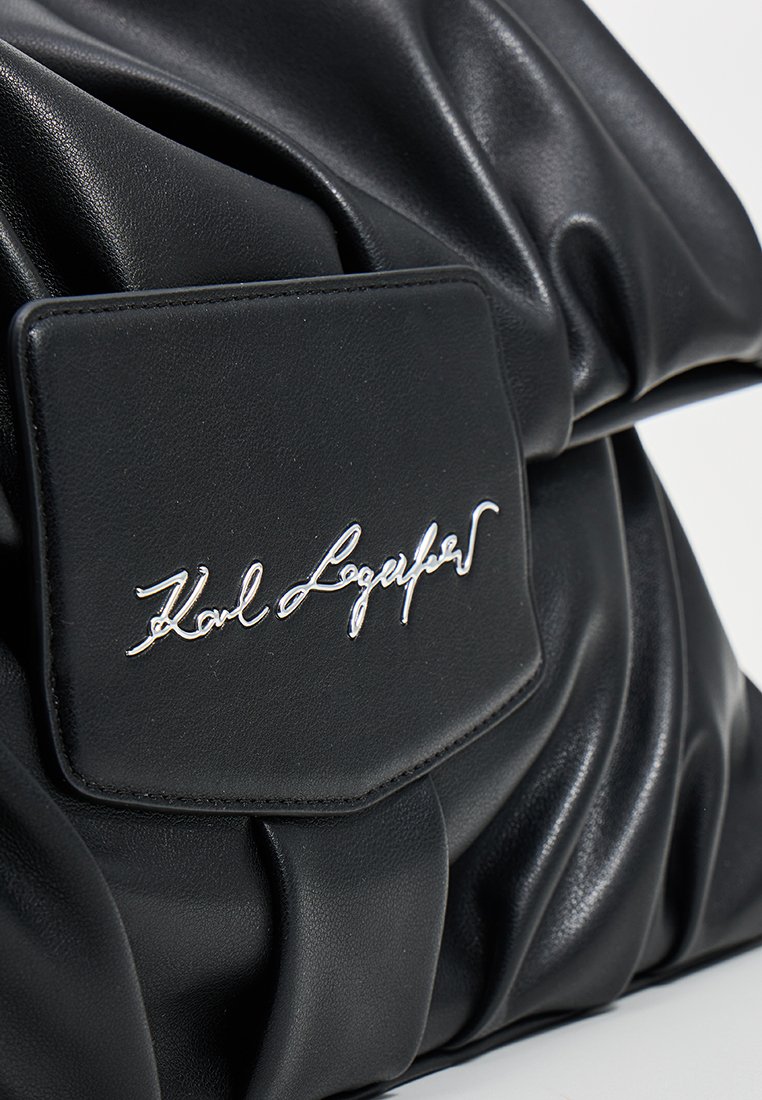 Sac en cuir noir avec un design froncé, doté d'une grande étiquette logo en métal argenté et d'une texture lisse. Apparence minimaliste et contemporaine.