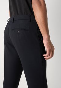 Homme portant un pantalon noir sur mesure avec poche arrière à bouton et passants de ceinture, montré de la taille à la cuisse avec la main détendue sur le côté.
