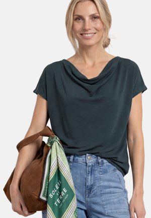 Uśmiechnięta kobieta w ciemnej bluzce z marszczonym dekoltem i jeansach z wysokim stanem, trzymająca brązową torbę z zielonym szalikiem przywiąznym do uchwytu.