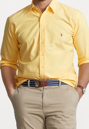 Mężczyzna w żółtej koszuli na guziki z podwiniętymi rękawami, beżowych spodniach, niebieskim pasku w paski i zegarku na nadgarstku, z rękami w kieszeniach.