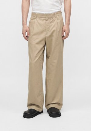Homme portant un pantalon beige ample et des chaussures noires, debout devant un fond clair uni.