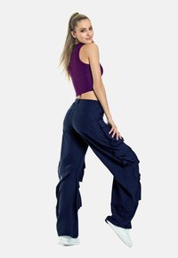 Lila kortärmad crop-top i kombination med marinblå cargobyxor med sidofickor och avslappnad passform. Modellen står med en hand på höften.