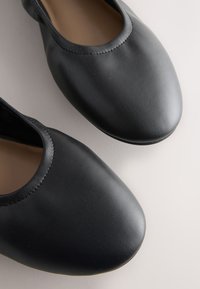Paire de ballerines plates en cuir noir à enfiler avec bouts arrondis sur fond clair.