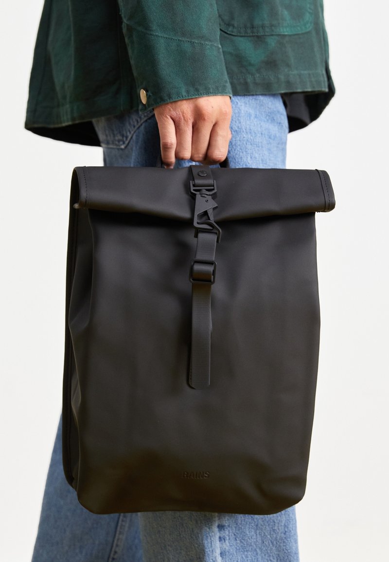 Personne tenant un sac à dos noir à rabat roulé avec boucle, portant une veste verte et un jean bleu sur un fond clair uni.