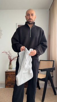 Mees, kellel on habemega paljas pea, kannab musta dressipluusi ja hoiab heledat halli kapuutsi heledas toas, kus on tool ja puidust kapp.