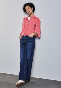 Camisa de pana color coral con mangas remangadas, combinada con unos vaqueros rectos de color azul oscuro. El modelo se encuentra de pie frente a una pared gris claro.
