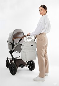 Grå klapvogn med en stofkappe og trækant i træ, suppleret af en hvid quiltet pusletaske. Model i beige bukser og hvid skjorte.