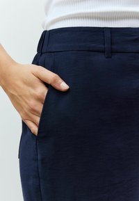 Tamno plave hlače s glatkom teksturom, s bočnom džepom i ravnim pojasom. Ruka opušteno položena u džepu.
