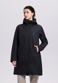 Mujer con impermeable negro con capucha y manos en los bolsillos, mirando ligeramente hacia un lado contra un fondo claro y sencillo.