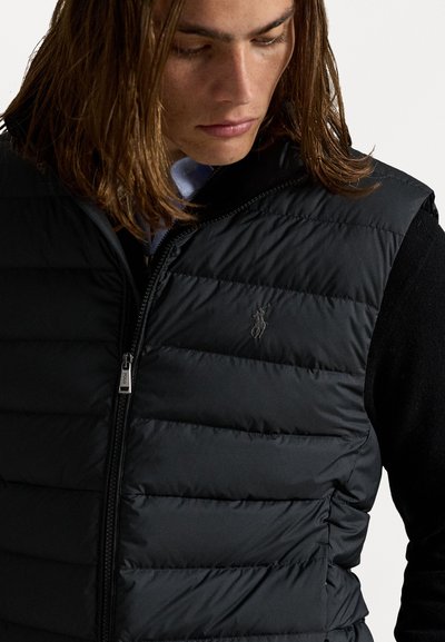 Sort puffer vest med horisontale quiltede sektioner og lynlås foran. Har et lille, præget logo på brystet. Bæres over en sort trøje.