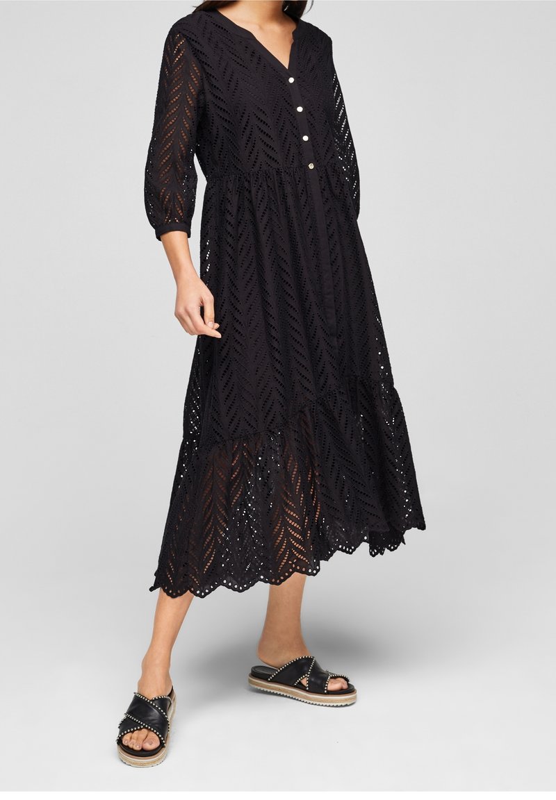 Femme portant une robe midi noire avec motif en dentelle, manches trois-quarts, boutons blancs et sandales noires cloutées à semelles compensées.