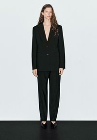 Black tailored suit featuring a deep V-neck blazer with two buttons, straight-leg trousers, and pointed toe black shoes on a light background.