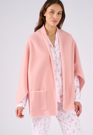 Femme portant un pyjama blanc à motifs floraux et un châle rose doux avec des poches avant, debout devant un fond clair uni.