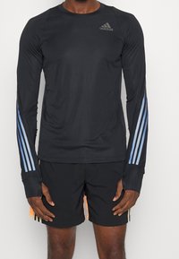 Camiseta deportiva negra de manga larga con acentos de tres rayas azules en las mangas, que presenta un cuello redondo y un diseño elegante y ligero.