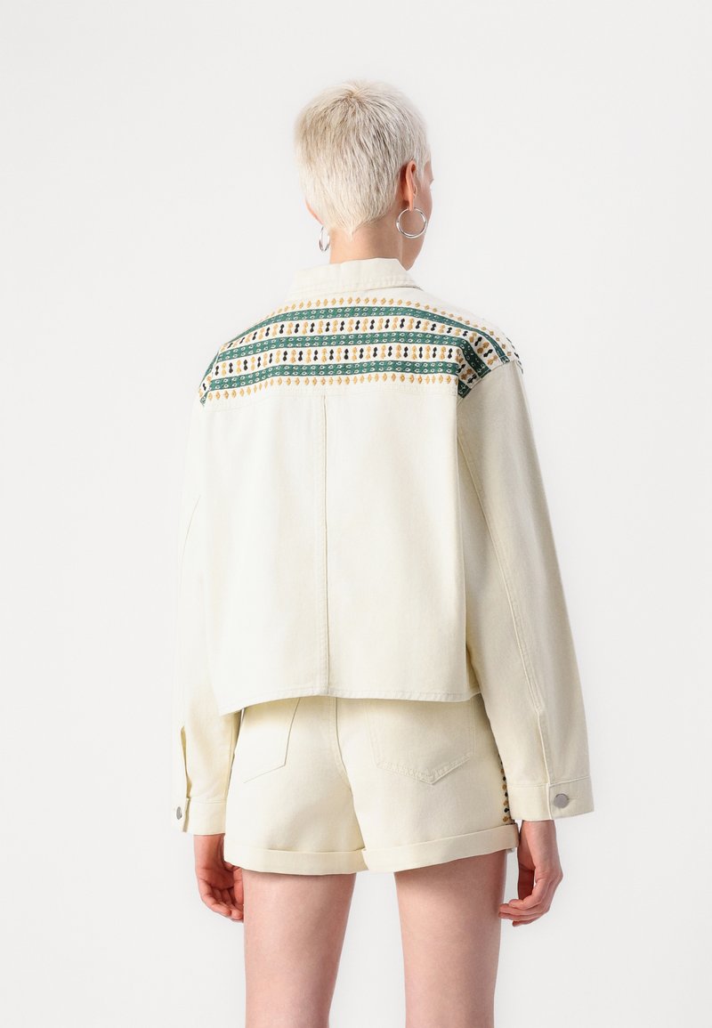 Veste en jean beige clair avec un design cropped. Elle présente des motifs brodés colorés au dos. Le tissu est doux avec une coupe décontractée.