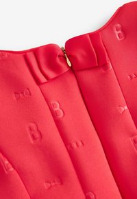 Détail d'une robe rouge mettant en avant un tissu lisse avec un motif en relief en forme de "B", un col arrondi, et une fermeture éclair ornée d'un accent en forme de nœud.