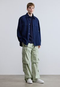 Πουκάμισο navy denim με δύο τσέπες στο στήθος, συνδυασμένο με ανοιχτό πράσινο cargo παντελόνι με πλαϊνές τσέπες, φορεμένο με λευκά sneakers.