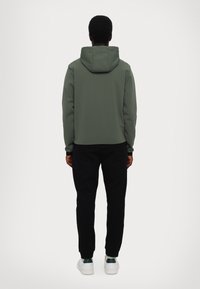 Giacca a vento verde oliva con un texture liscia e cappuccio regolabile; abbinata a pantaloni neri a gamba affusolata e sneakers bianche. Design minimalista.