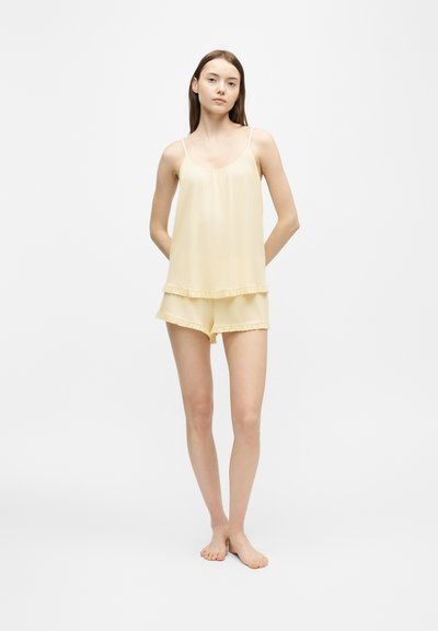Jeune femme debout pieds nus portant un haut sans manches jaune clair et un short assorti avec des bords à volants sur un fond uni.