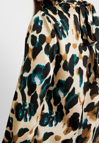 Farklı desenli bluz, saten bir görünüm sunuyor, sıcak bej bir zemin üzerinde cesur teal, siyah ve kahverengi benekler var; yakada bağlama detayı ile birlikte.