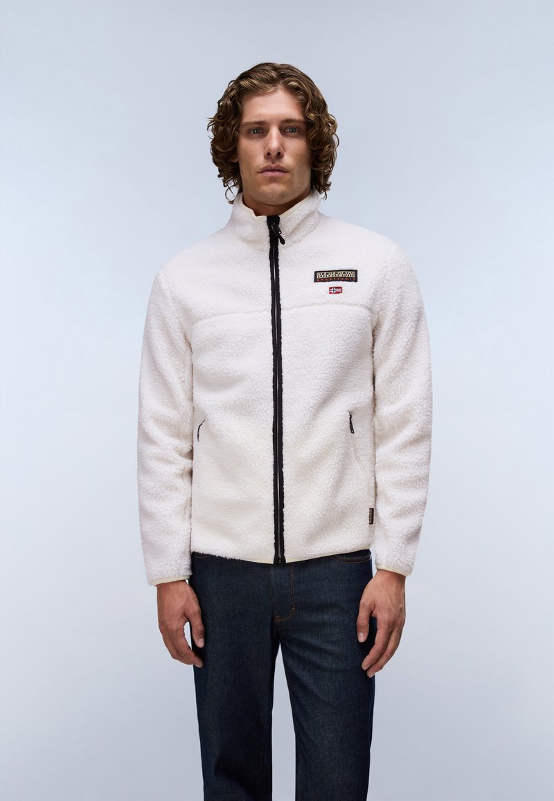 Mann mit lockigem Haar, der eine weiße Fleecejacke mit Reißverschluss und dunkle Jeans trägt, steht vor einem einfarbigen hellgrauen Hintergrund.