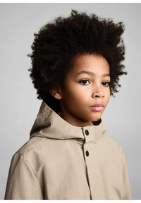 Khaki kapucar jakna iz gladke tkanine s črnimi deli; model ima kodrasto temno lase in nevtralen izraz na sivi ozadju.