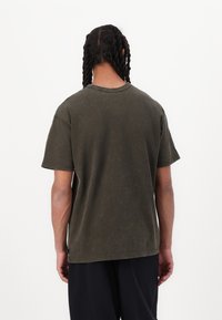 Tricou din bumbac de culoare verde măslin, cu o croială lejeră, mâneci scurte și gât rotund. Țesătura are o textură subtilă și un aspect uzat.