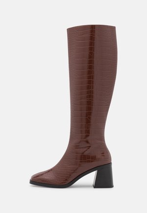 Botte haute marron jusqu'au genou avec motif crocodile, bout pointu et talon moyen noir épais sur fond clair uni.