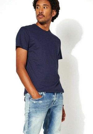 Man met krullend haar die een donkerblauw T-shirt en vervaagde blauwe jeans draagt, kijkt opzij met één hand in de zak.