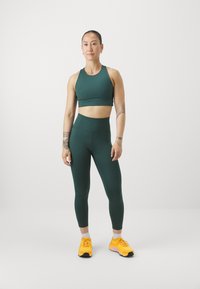 Zielony stanik sportowy i legginsy z wysokim stanem, o gładkiej strukturze. Noszone z jasnoniebieskimi butami sportowymi. Proste tło.