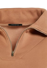 Acercamiento de un cuello con cremallera marrón en una prenda, con un tirador de cremallera plateado parcialmente abierto, mostrando la suave textura de la tela.
