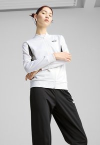 Bijela zip-up jakna s crnim bočnim panelima, napravljena od glatkog materijala, uparena s crnim hlačama. Jakna ima Puma logo na prsima.