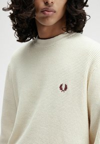 Cremefarbener Waffelstrickpullover mit hohem Kragen und maroonfarbigem aufgesticktem Logo auf der Brust. Weiche Textur und lockere Passform.