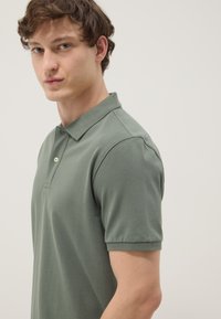 Joven con cabello corto, rizado y castaño, que lleva una camiseta polo de manga corta de color verde claro, mirando ligeramente hacia la izquierda sobre un fondo liso.