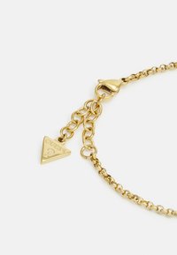 Bracelet en chaîne dorée avec fermoir, comportant un pendentif triangulaire gravé avec "GUESS". Texture lisse et maillons de chaîne intriqués.
